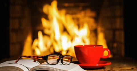 Table holding book, reading glasses, and mug in front of burning fireplace