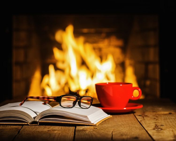 Table holding book, reading glasses, and mug in front of burning fireplace