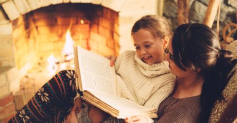 Mother reading book to daughter in front of a warm fireplace