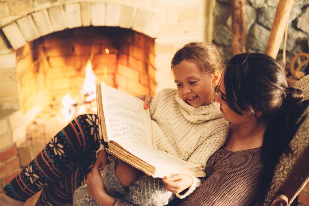 Mother reading book to daughter in front of a warm fireplace