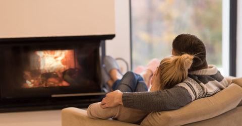 Young romantic couple sitting on sofa in front of fireplace at home, looking at each other, talking and drinking coffee at autumn day