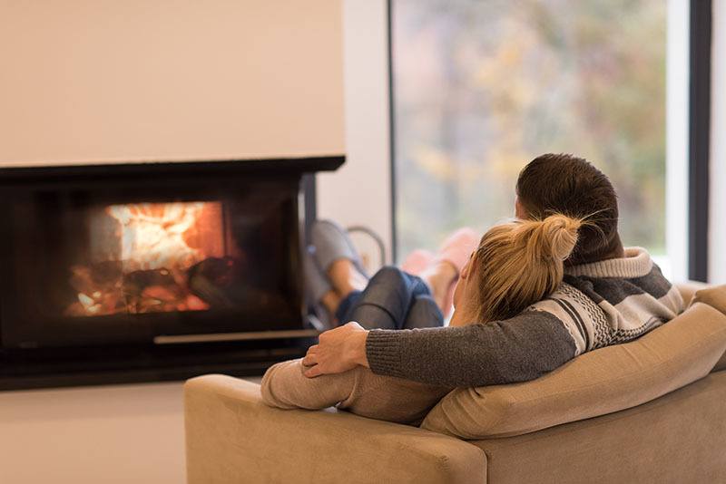 Young romantic couple sitting on sofa in front of fireplace at home, looking at each other, talking and drinking coffee at autumn day