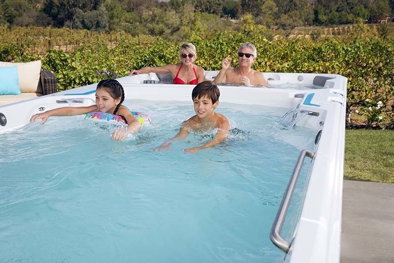 a family of two young children and a man a women enjoying swimming in a swim spa outside on a sunny day