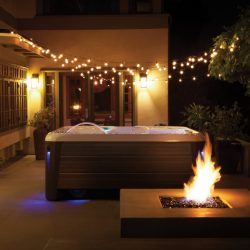 Spa on patio at night with string lights overhead and a lit fire pit in the foreground