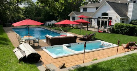 Lush green backyard with lawn, bushes and trees, and a full swim spa with deck and pool next to a white home.