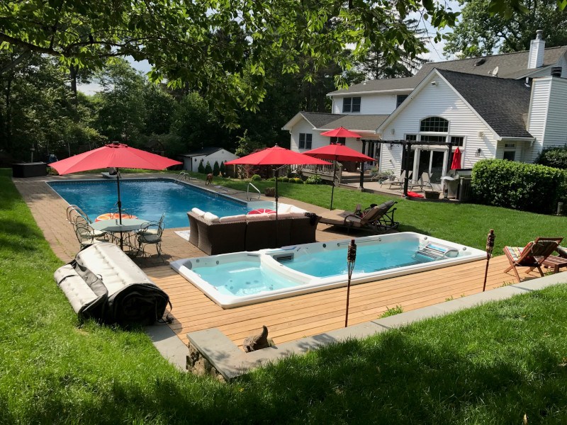 Lush green backyard with lawn, bushes and trees, and a full swim spa with deck and pool next to a white home.