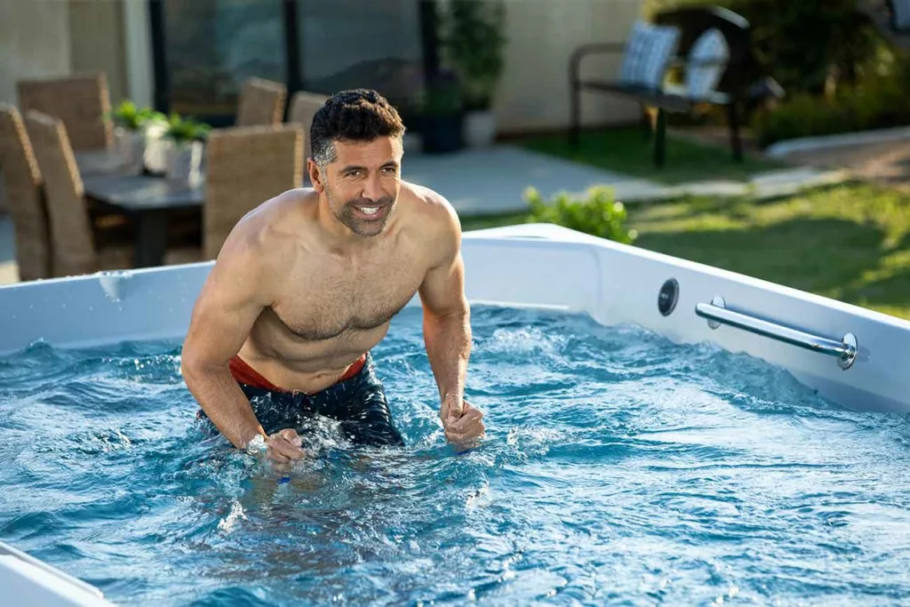 Muscular man with graying temples smiling while using Endless Pools aquabike in backyard swim spa.