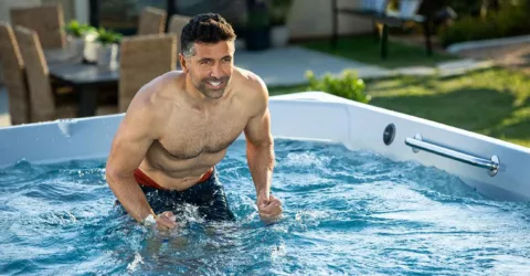 Muscular man with graying temples smiling while using Endless Pools aquabike in backyard swim spa.