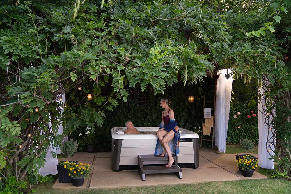 Man in spa and woman on edge with feet on steps. Spa sits on patio under a canopy of greenery and has open privacy curtains on two ends. 