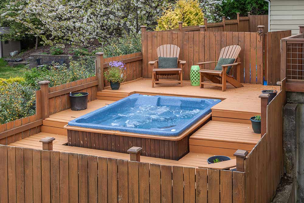 Hot tub with blue shell built into a tri-level deck in a backyard. Two Adirondack chairs and potted plants also sit on deck.  