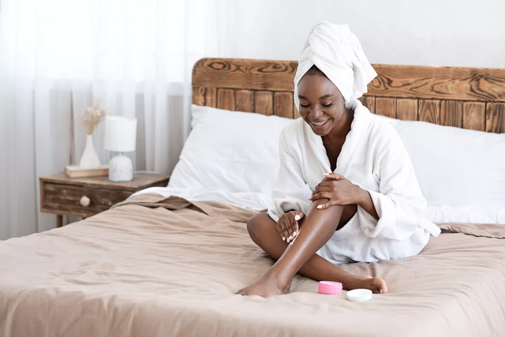 Young woman in bathrobe with towel around her head puts lotion on her legs while sitting on bed. 