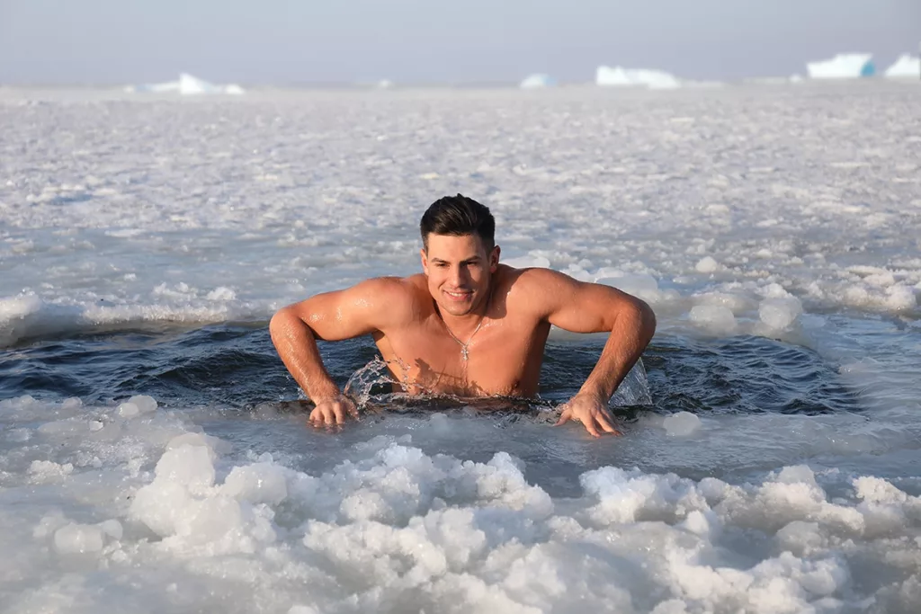 Young athletic man in bathing suit participating in cold plunge Cryotherapy in a partially frozen body of water for the medicinal health benefits.