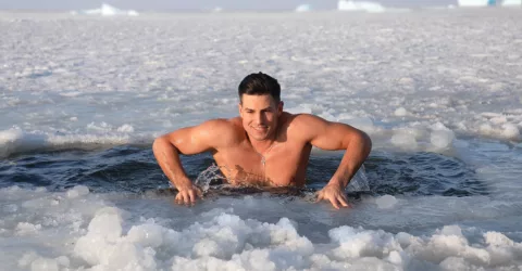 Young athletic man in bathing suit participating in cold plunge Cryotherapy in a partially frozen body of water for the medicinal health benefits.