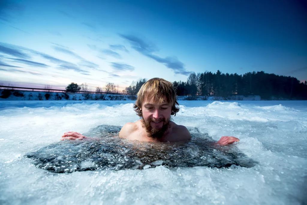 A man with a beard takes a cold plunge in an icy lake surrounded by snow.