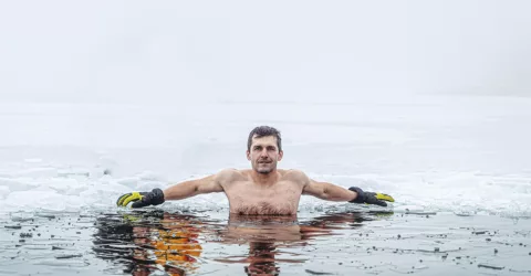 An athlete takes a cold plunge in an ice hole surrounded by snow