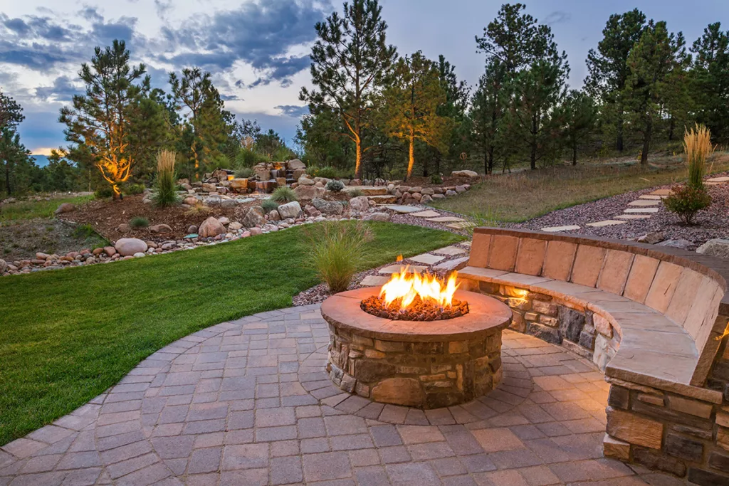 A stone firepit in a backyard in the evening, surrounded by trees and green grass.
