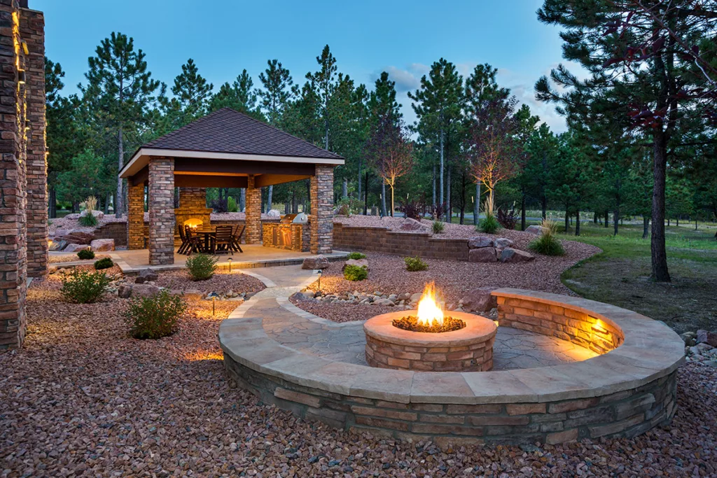A firepit at night in a spacious backyard with outdoor seating and trees.