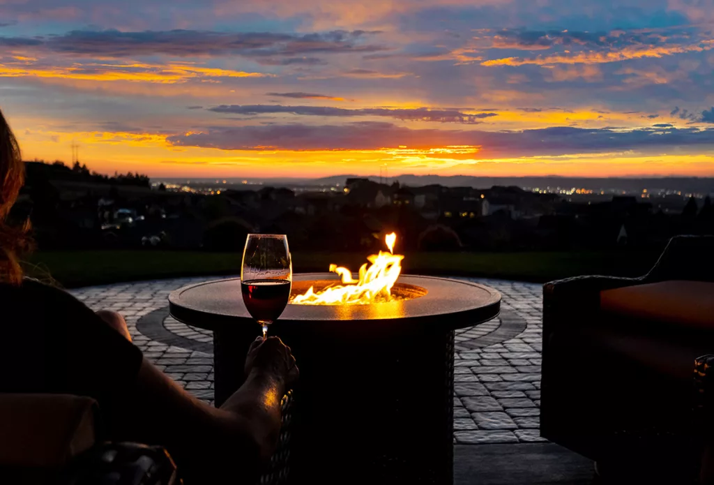 A woman drinks a glass of wine in front of a firepit at sunset