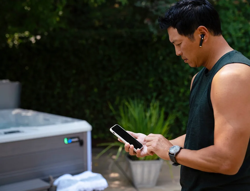 A man standing next to his hot tub looking down at his smartphone, using the latest smart spa technology for hot tubs, an app to check to see if his hot tub water is balanced