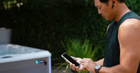 A man standing next to his hot tub looking down at his smartphone, using the latest smart spa technology for hot tubs, an app to check to see if his hot tub water is balanced