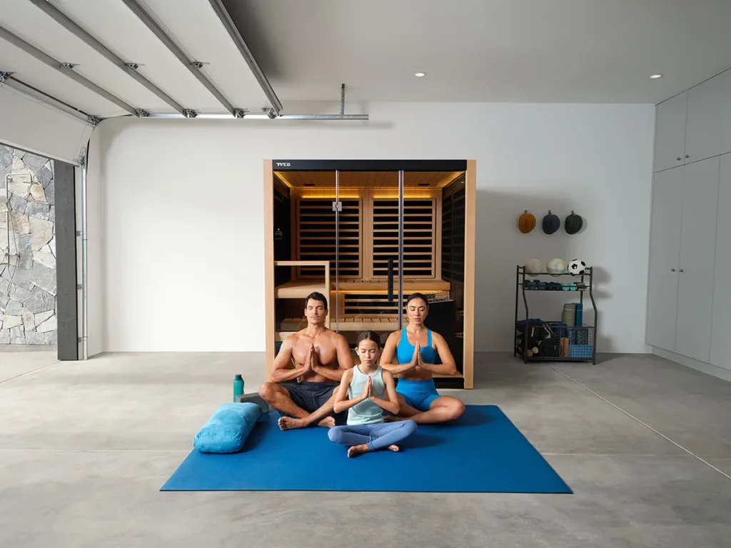 A family of three sits on a blue yoga mat in the garage, meditating in front of an infrared sauna