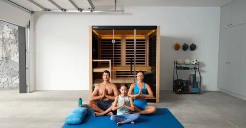 A family of three sits on a blue yoga mat in the garage, meditating in front of an infrared sauna