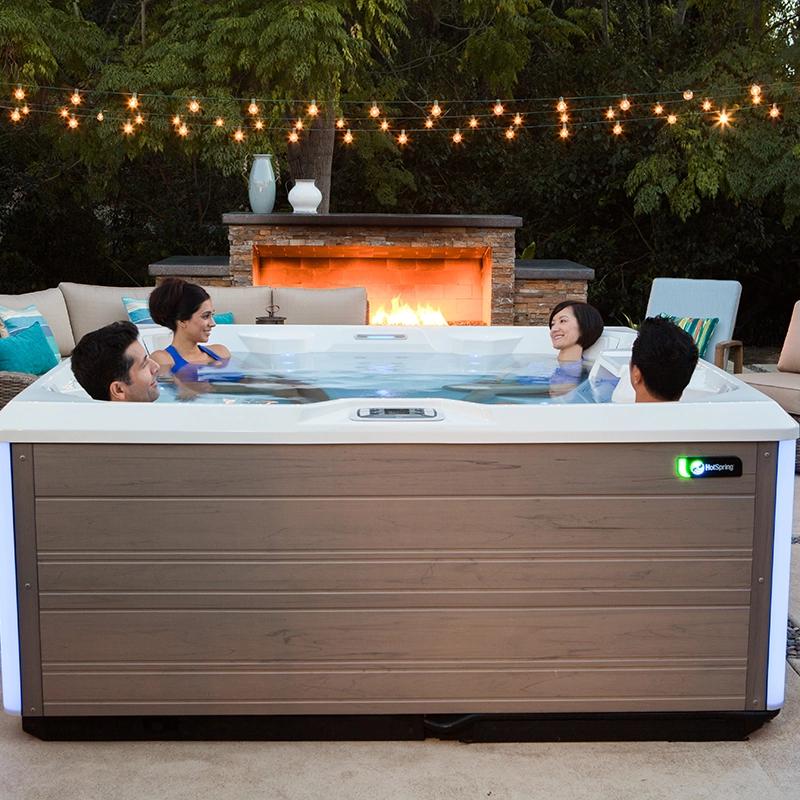 Four smiling adults relaxing in an outdoor Hot Springs hot tub, surrounded by trees and fairy lights