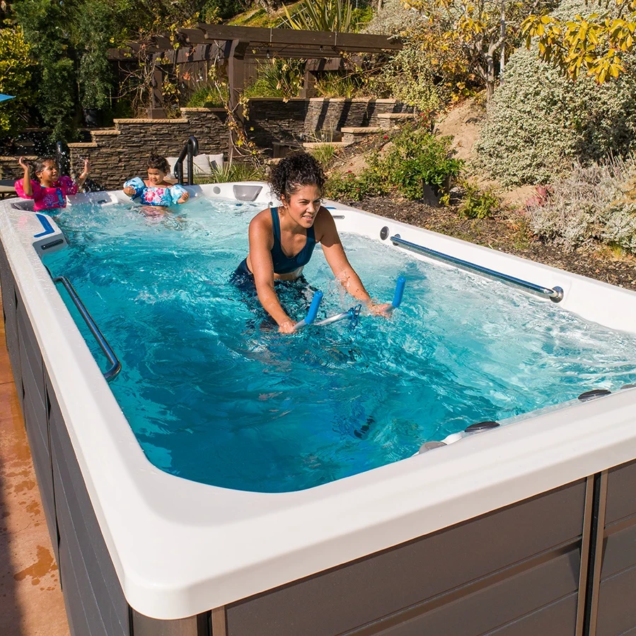 A women exercising on an aquatic bike in an X-Series SwimCross Swim spa, while her 2 young daughters play in the water near her.