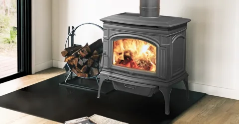 A cast iron wood burning stove in a living room with hardwood floors and white walls, next to a stack of firewood and a sliding glass door