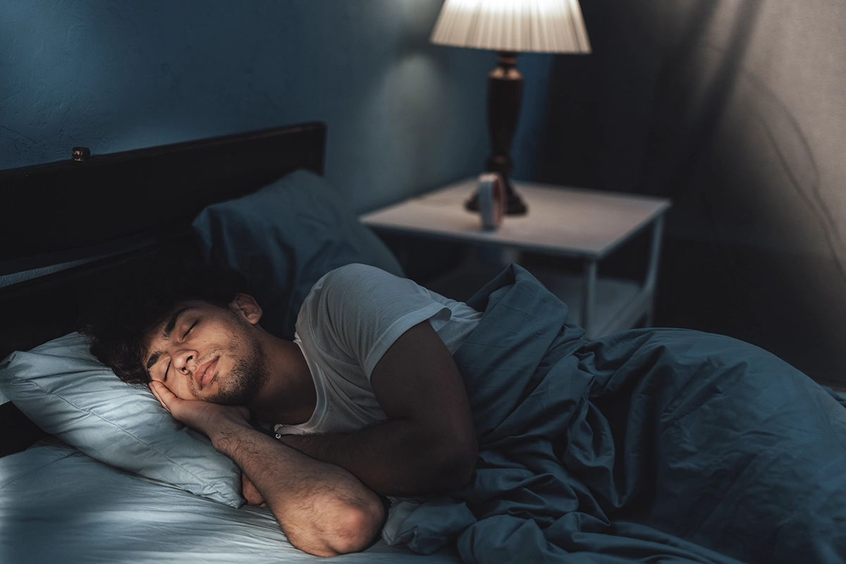 A young man sleeps peacefully in bed at night time, with blue lighting and a lamp on the nightstand, showing the concept of better sleep